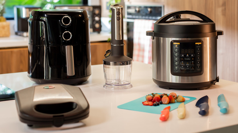 Variety of kitchen appliances, knives, and a cutting board with fruit on a kitchen counter