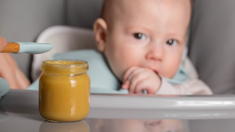 a 5-6 mo old baby in a highchair looking at a jar of baby food on his tray