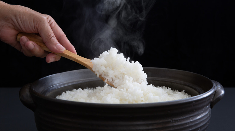 Steaming hot cooked white rice being spooned from a black clay pot