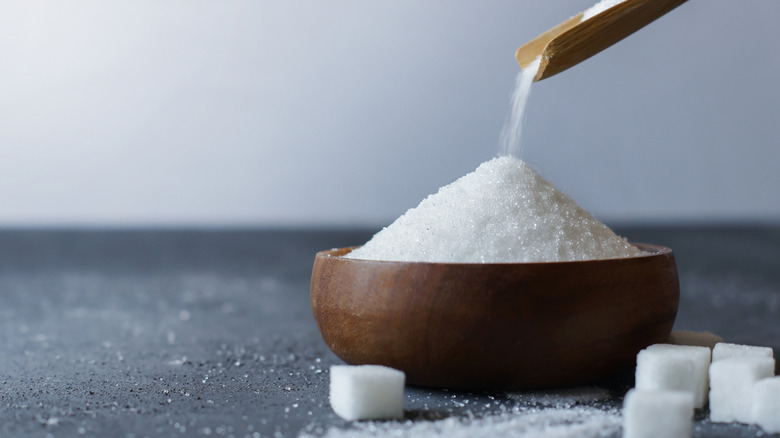 wooden spoon pouring granulated sugar into a wooden bowl with sugar cubes in foreground