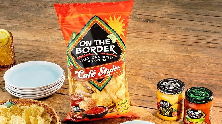a packet of On The Border tortilla chips with a stack of plates, a plate of chips and 2 jars with dips on the side