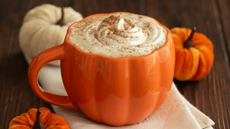 Orange, pumpkin-shaped mug of coffee, whipped cream, and spices surrounded by plush pumpkins