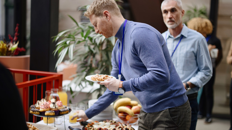 A man takes some buffet food while another man waits behind him