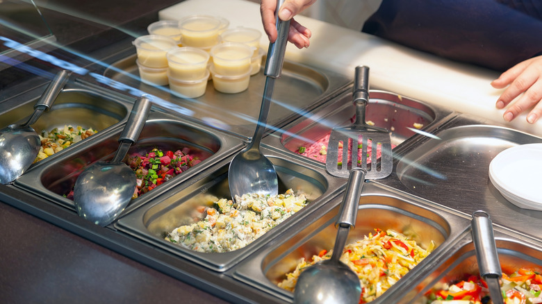 A hand holds a spoon at a buffet, which is sitting in a salad