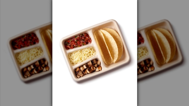 A Chipotle take out box with two hard taco shells, chicken, cheese, and pico de gallo against a white background