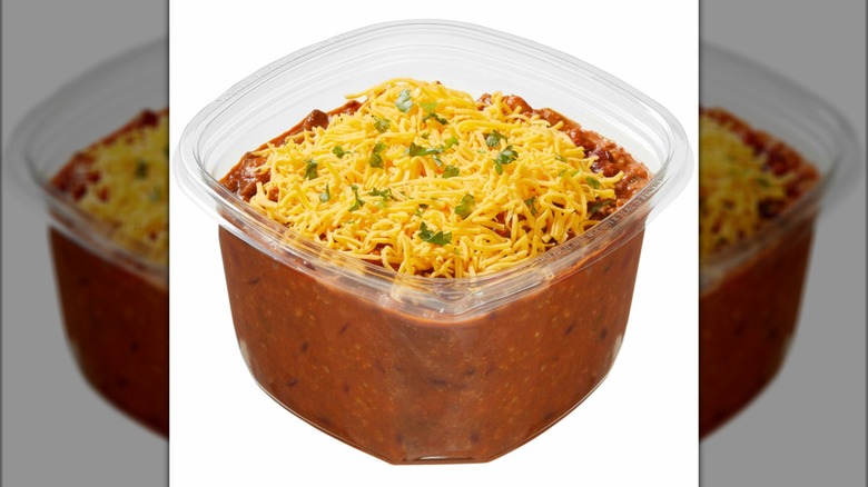 A plastic container of beef chili with shredded cheddar cheese on top on a white background
