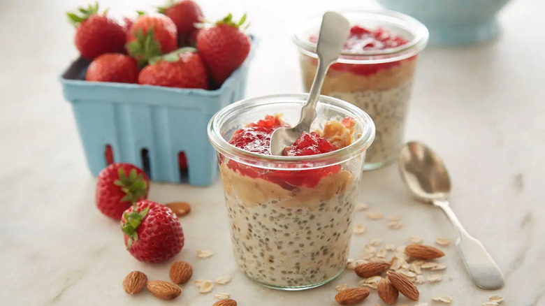 Two cups of overnight oats with almond butter and jam on the top on a table with a basket of strawberries and some scattered almonds