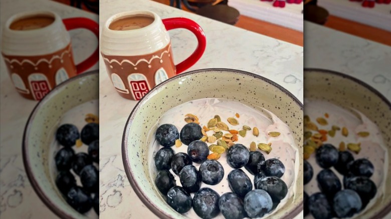 A bowl of Greek yogurt with blueberries and pumpkin seeds on a counter with a cup of coffee in the background