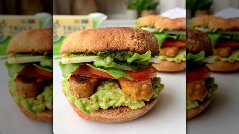 Closeup of a sandwich with English muffin, tempeh, tomato, avocado, and greens on a counter