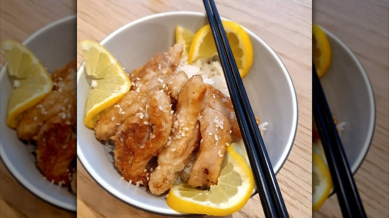 A bowl full of lemon chicken with lemon slices, rice, and chopsticks on a table
