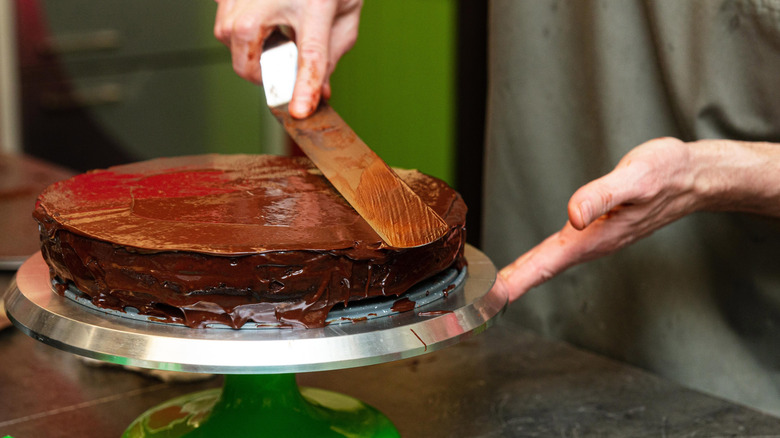 A baker spreading frosting on a chocolate cake placed on a turntable with an offset spatula