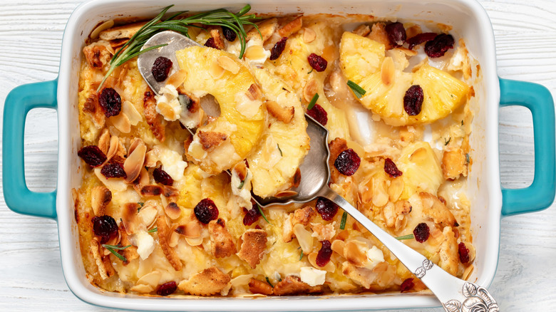 overhead shot of a serving spoon digging into a pineapple casserole topped with sliced almonds and dried cranberries
