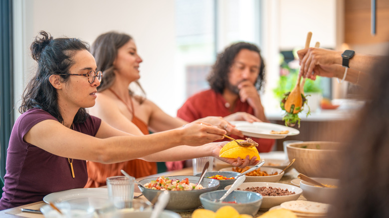 A group of friends eat at a dinner table, with one person preparing a taco, and others passing around food