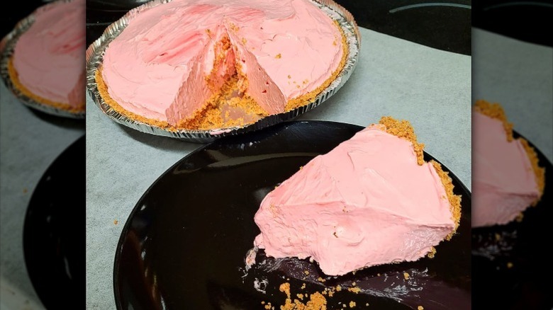 A slice of Kool-Aid pie on a plate with a Kool-Aid pie in an aluminum tin placed on a table