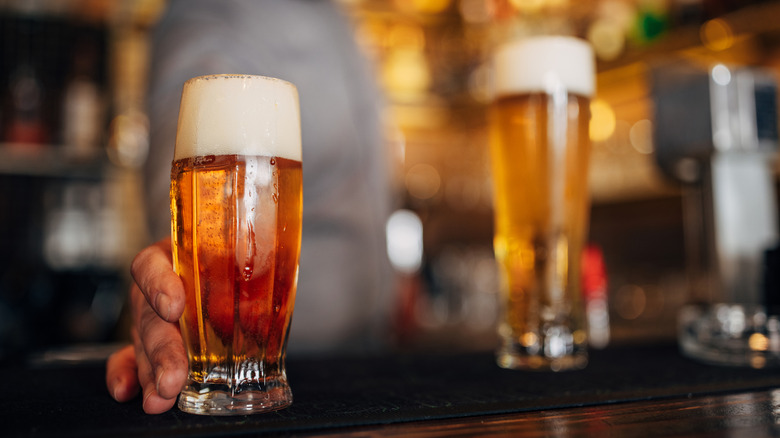 A bartender holds a glass of beer at a bar, with another beer out of focus in the background