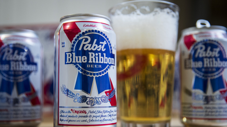 Cans of Pabst Blue Ribbon are seen next to a glass of half-drunk beer