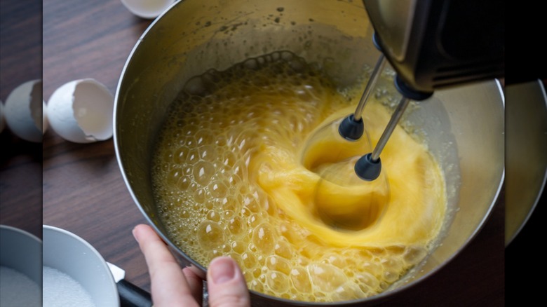 A person whipping eggs with sugar in a bowl using a hand mixer