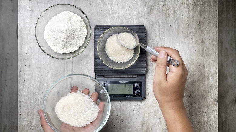 A person weighing out sugar in a bowl on a kitchen scale with a bowl of flour on the side
