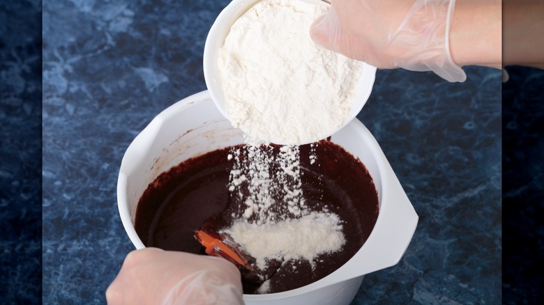 A person adding flour to a white bowl full of brownie batter