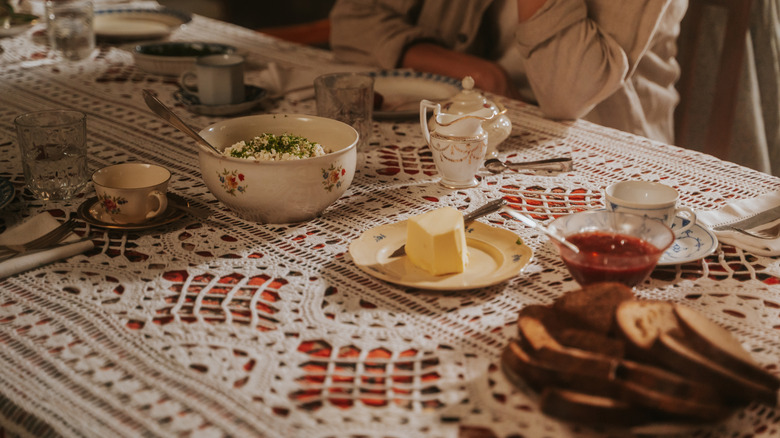 Person sitting at dining table with lace tablecloth, plus bread and butter, dishes.