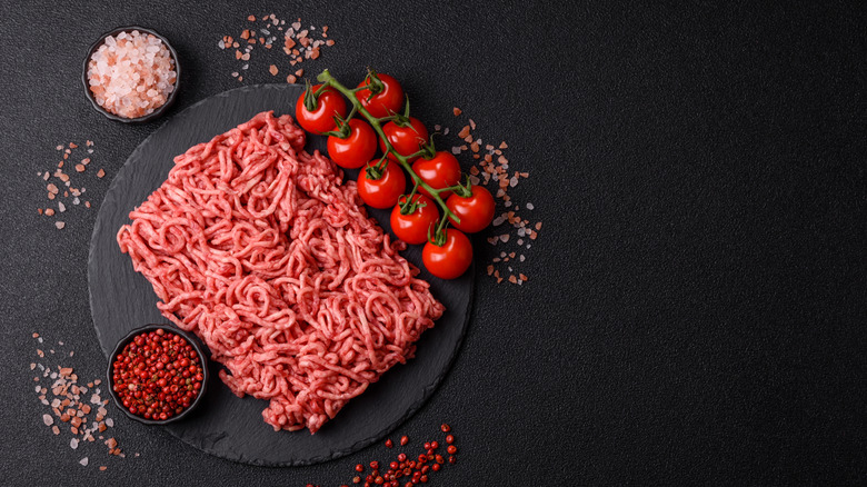 Ground meat, tomatoes, and bowls of pink salt and pink peppercorns on a black table