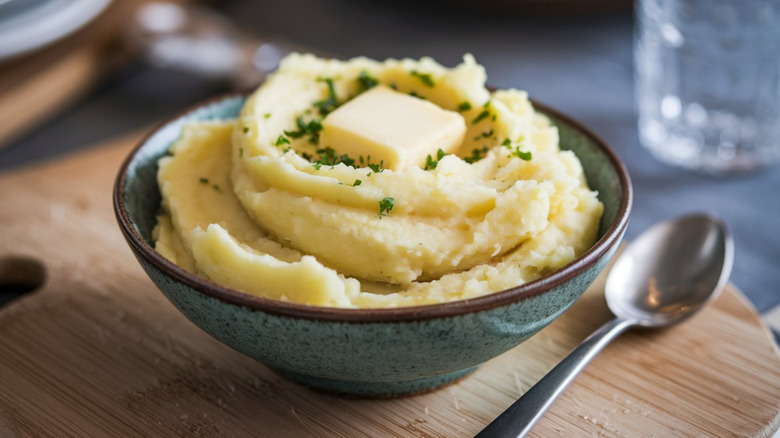 A ceramic bowl full of fluffy mashed potatoes topped with butter on a wooden board with a spoon