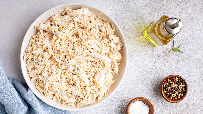 Shredded cooked chicken in a white bowl placed on a marble table with oil, salt, and pepper on the side