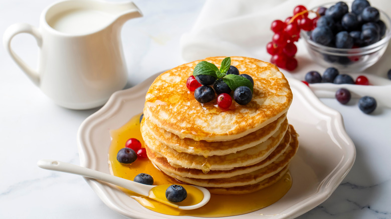 A stack of pancakes on a white plate with fresh fruit and honey next to a pitcher of milk