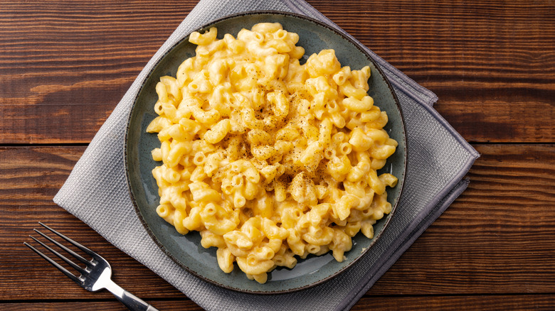 A plate full of mac and cheese placed on a napkin with a fork on the side, all on a wooden table