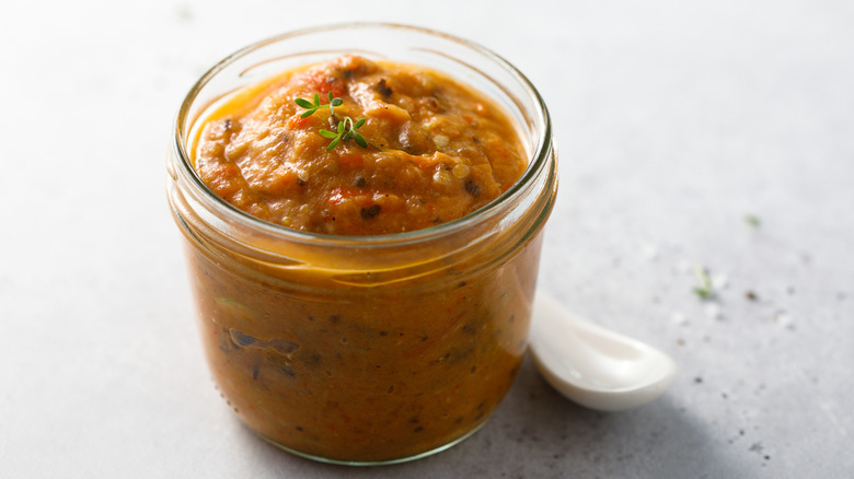 Pumpkin baba ghanoush in a glass jar with a spoon on the side, all on a white table