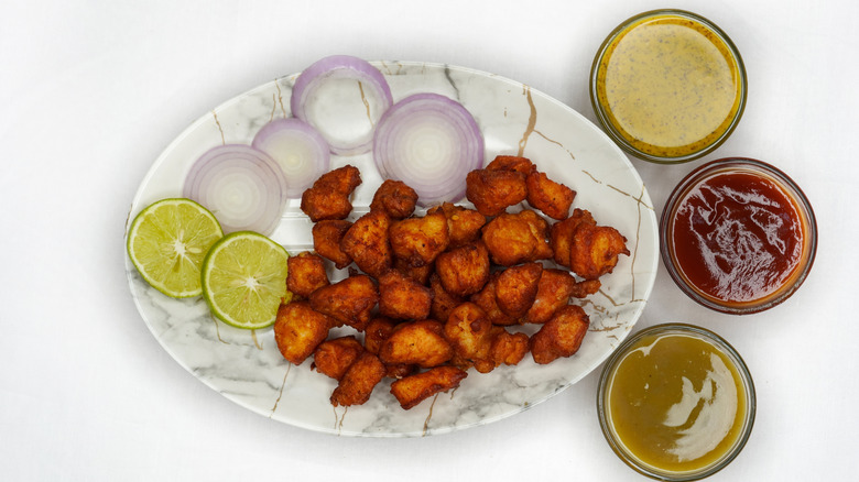 Chicken pakoras, lemon slices, and onion slices on a white plate, with three different types of dips on the side