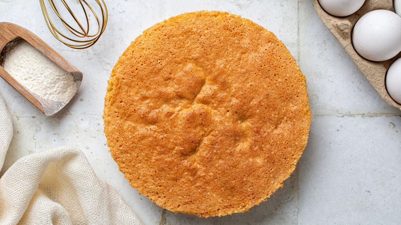 A whole vanilla cake on a marble table with eggs and flour on the side
