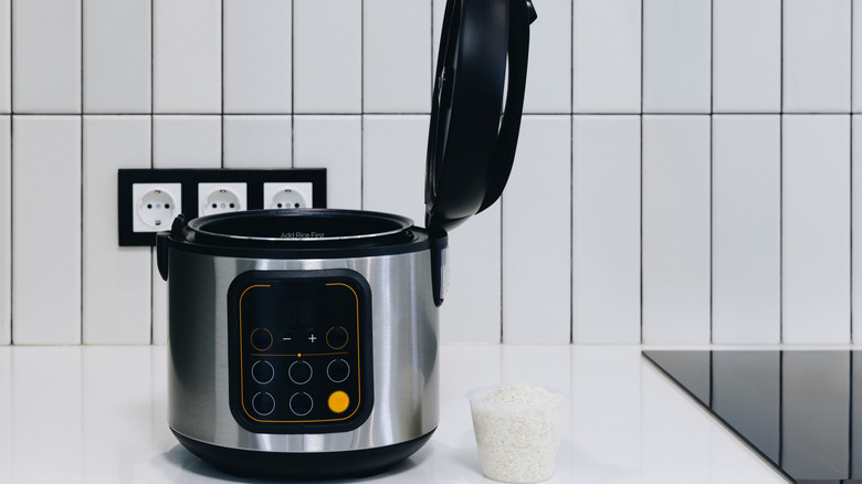 A rice cooker on a kitchen counter with a bowl of uncooked rice on the side