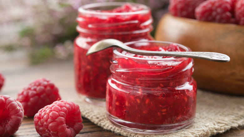 Two jars full of raspberry jam on a wooden table with fresh raspberries on the side