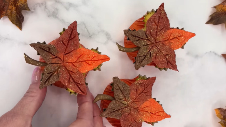 A hand placing a cupcake with a maple leaf made with frosting next to two others.