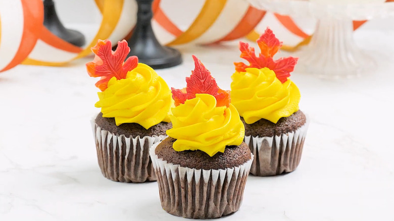 Three cupcakes with yellow frosting and orange candy maple leaves