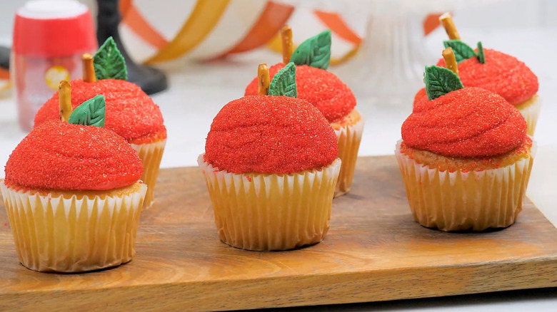 Six shiny apple cupcakes with sparkle frosting and green fondant leaves on a cutting board