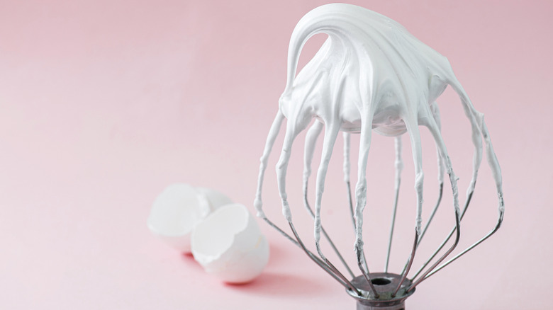 Freshly whipped Italian Meringue on a whisk placed with egg shells in the background