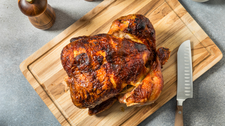 A rotisserie chicken sits on a wooden board, next to a knife