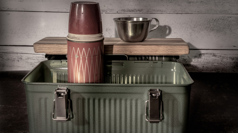 A vintage thermos and metal cup sticking out of an open vintage steel lunch box