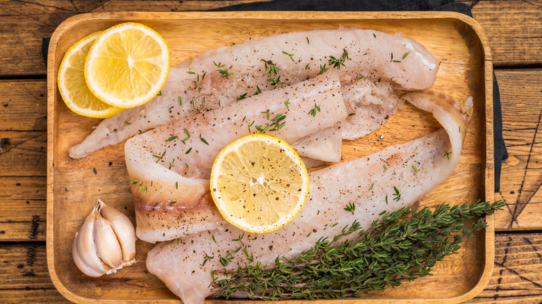Fish fillets on a wooden board with slices of lemon, garlic, and rosemary on the side