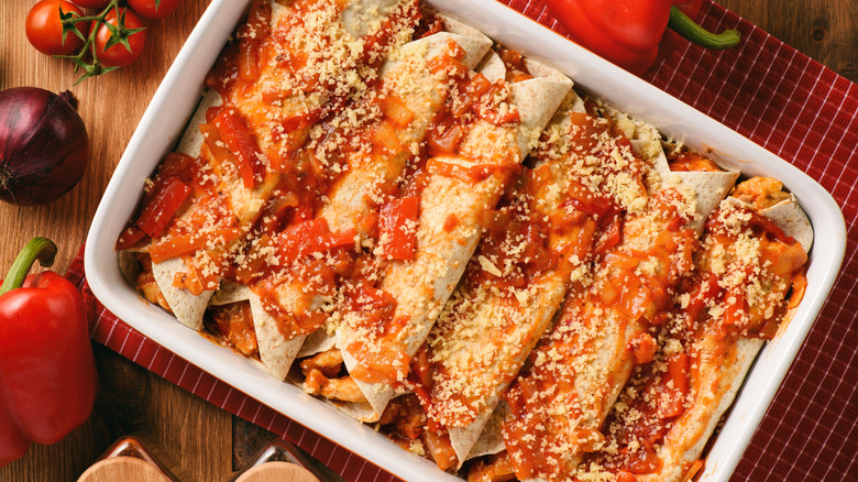 A tray of enchiladas with red sauce is seen from above, with fresh vegetables surrounding it