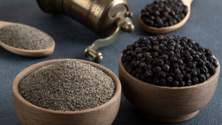 Wooden bowls of black peppercorns and ground black pepper, with a mill in the background.