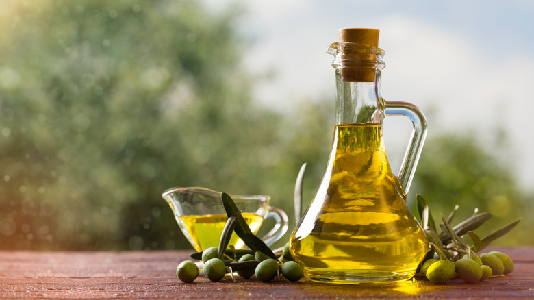A glass decanter of olive oil on a table next to some fresh olives still attached to the branches.