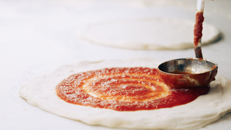 Pizza sauce is being evenly distributed across an uncooked pizza crust dough with a ladle