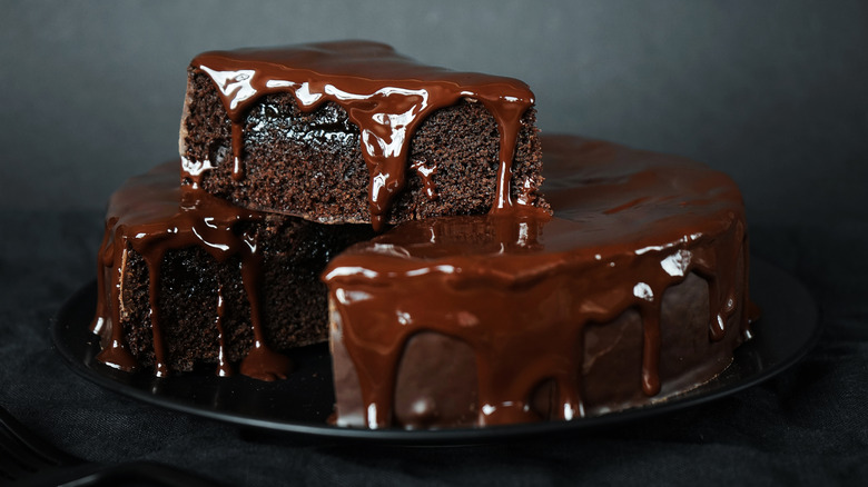 A chocolate cake topped with ganache with a slice removed perched atop the whole cake
