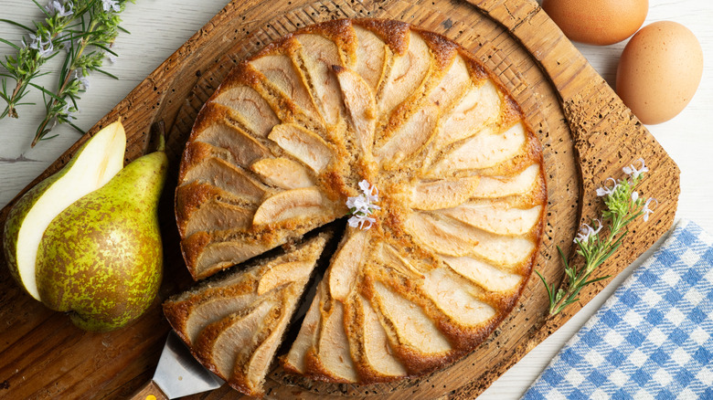 Pear cake with a slice being removed on a wooden cutting board garnished with a fresh pear and sprig of herb