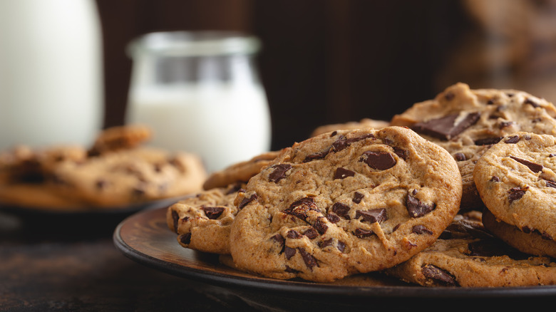 Freshly baked chocolate chip cookies on a black plate with a glass of milk in the background