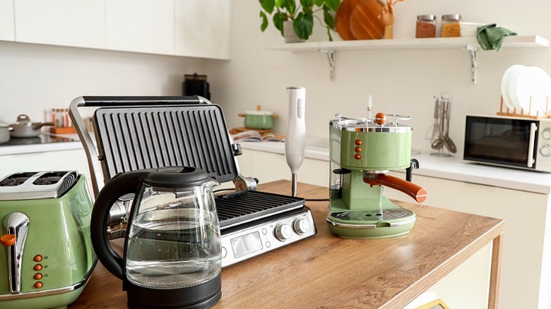 an open grill with a toaster, a water jug and a coffee maker on a kitchen top with the kitchen counter and more appliances int he background