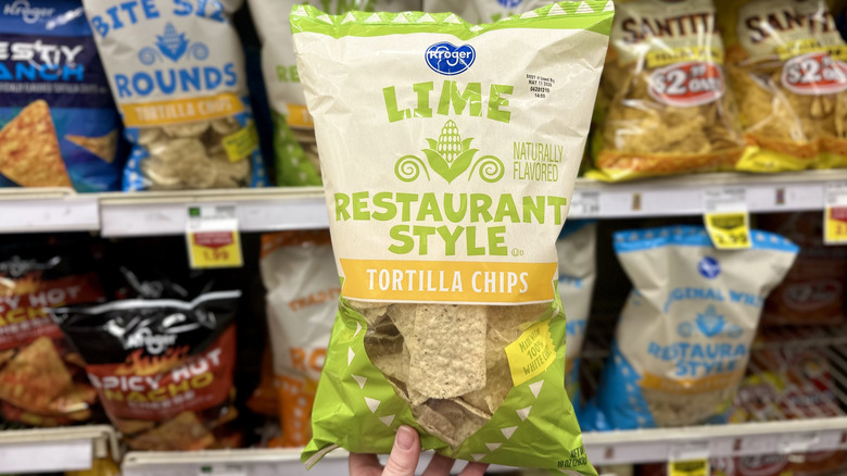 A Kroger Kick of Lime restaurant style corn tortilla chips bag in front of a shelf of other chips